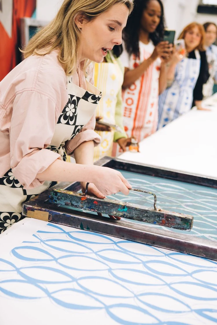 A woman in a pink shirt and patterned apron demonstrates screen printing to a group of workshop participants.