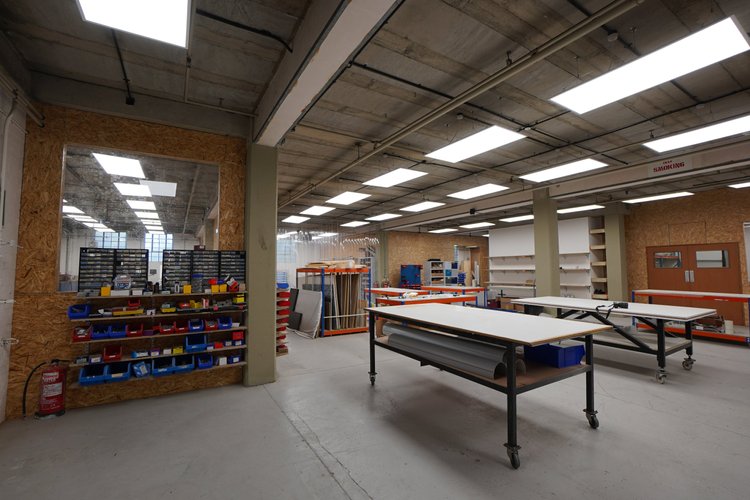 A spacious workshop or maker space with worktables, shelves, and organized storage bins, illuminated by overhead fluorescent lights.
