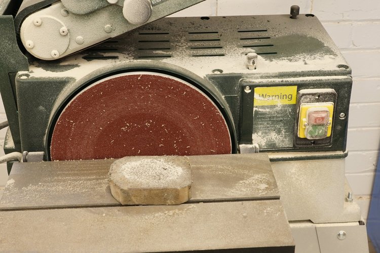 A sanding or polishing machine with a red abrasive wheel, covered in dust, and a piece of stone or material resting on the machine's surface, also dusted.