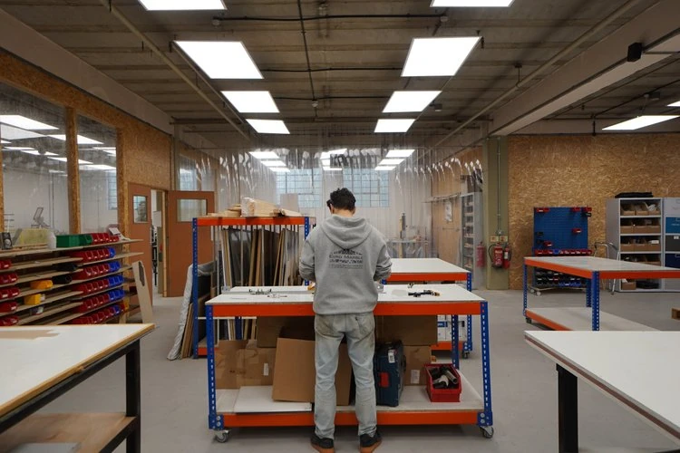 A person in a gray hoodie working at a workshop table with various tools and materials, in an industrial woodworking space with storage shelves, tools, and polycarbonate wall panels.