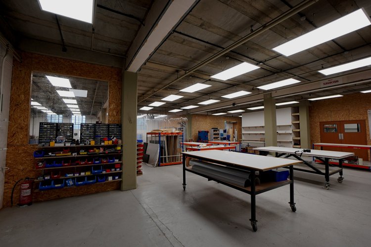 Open workshop with worktables and parts storage beneath exposed concrete beams.
