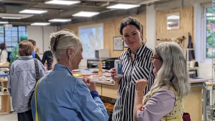 Three women talking and smiling at a craft workshop or classroom, with other people and tools in the background.