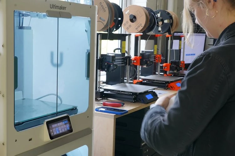 A person working with 3D printers in a workshop, with spools of filament overhead and a 3D printer in the foreground.