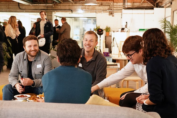 Group of five people sitting around a table, engaging in conversation and smiling at each other, during a social or professional gathering in an indoor venue with plants and background activity.