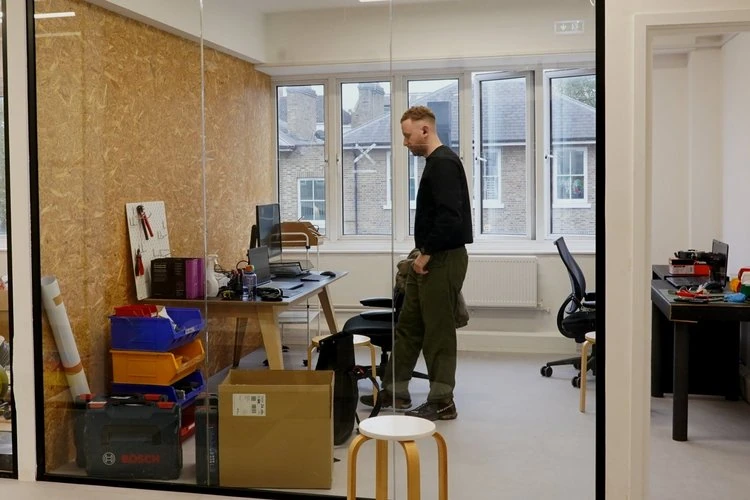 Man standing in an office room with desks, chairs, and tools, viewed through a glass wall