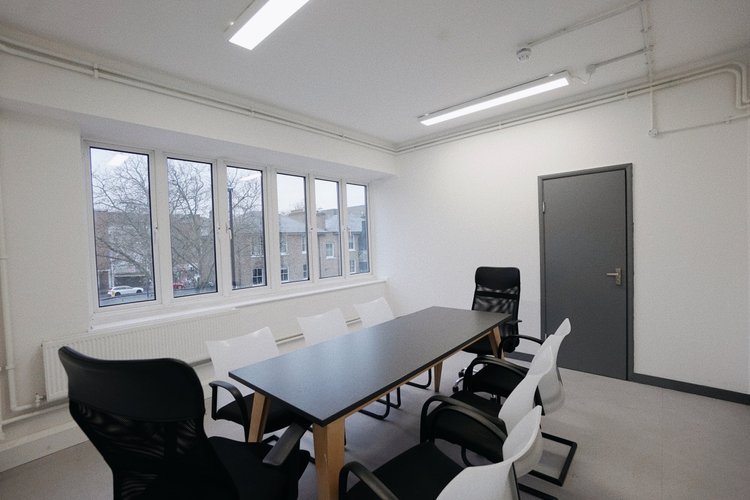 Empty conference room with six chairs, a black table, a window with a view of trees and buildings, white walls, gray door, and fluorescent lighting