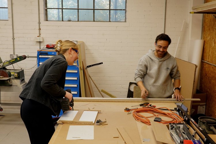 Two people working together on a woodworking project in a workshop, laughing and smiling.