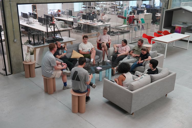 Group of people sitting and talking in a modern, open office lounge area with sofas, armchairs, a glass table, and a background of open workspace with desks and computers.