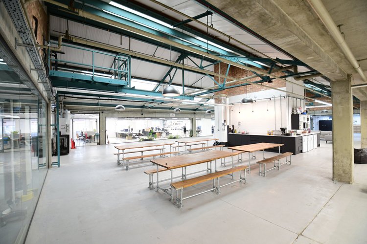 Spacious modern coworking space with wooden tables and benches, industrial design features, exposed pipes and beams, and a small kitchenette area.