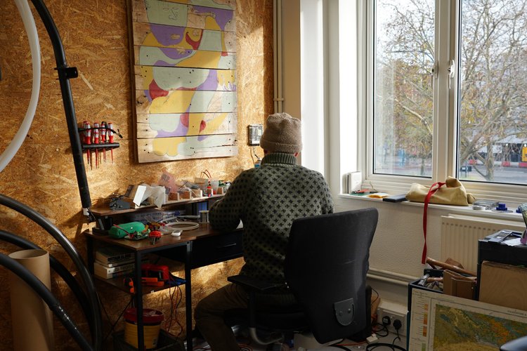 A person sitting at a cluttered desk in a room with a wooden wall and a large window, looking outside. There is abstract art on the wall and various electronic and craft materials scattered on the desk.
